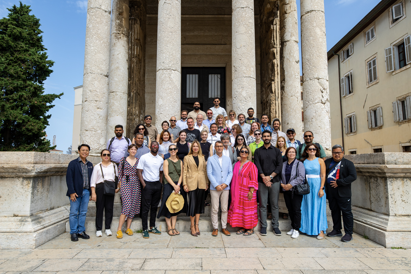 Group of professionals wearing various colours, smiling, standing on tiered steps going up to large columns in front of a hall.
