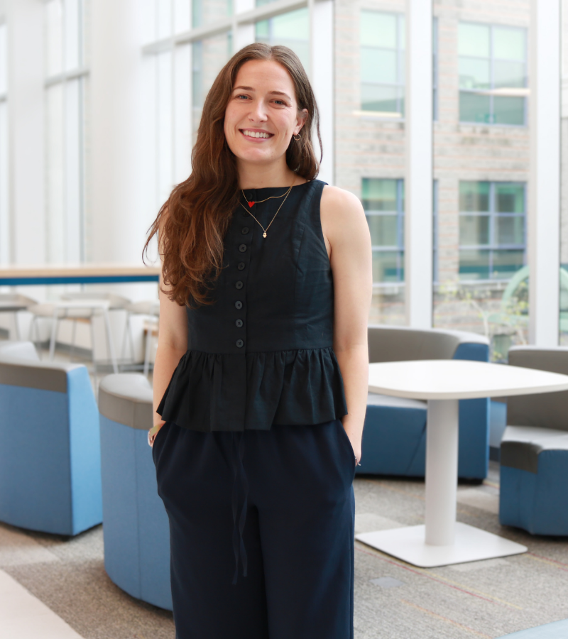 Rachel is a white woman with long brown hair. She wears a black top and pants and smiles with her hands in her pockets.